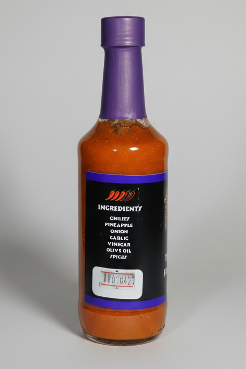 A bottle of Ingredients brand chilef pineapple onion garlic vinegar olive oil spice sauce, featuring orange-red sauce inside a clear glass bottle with a purple cap and black label.