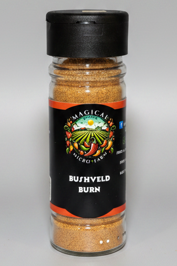 A clear plastic spice jar filled with brownish-orange seasoning powder, topped with a black cap. The front label is black with orange bands and a colorful farm logo, reading 'MAGICAL MICRO-TABBS BUSHVELD BURN'.