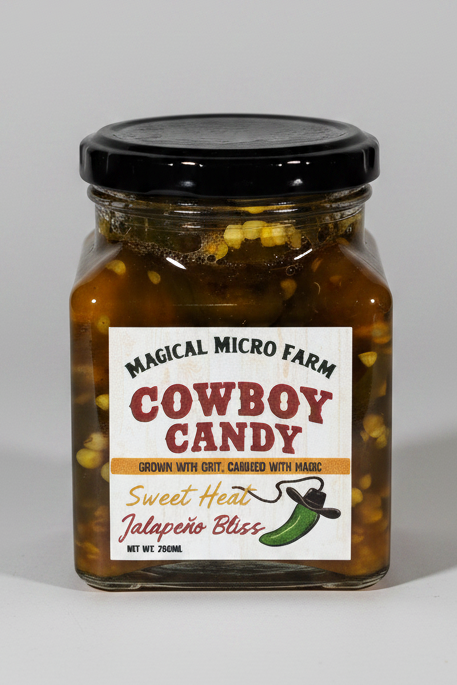 A glass jar of Magical Micro Farm Cowboy Candy Sweet Heat Jalapeño Blends candy with a black lid and a white label, isolated on a white background.