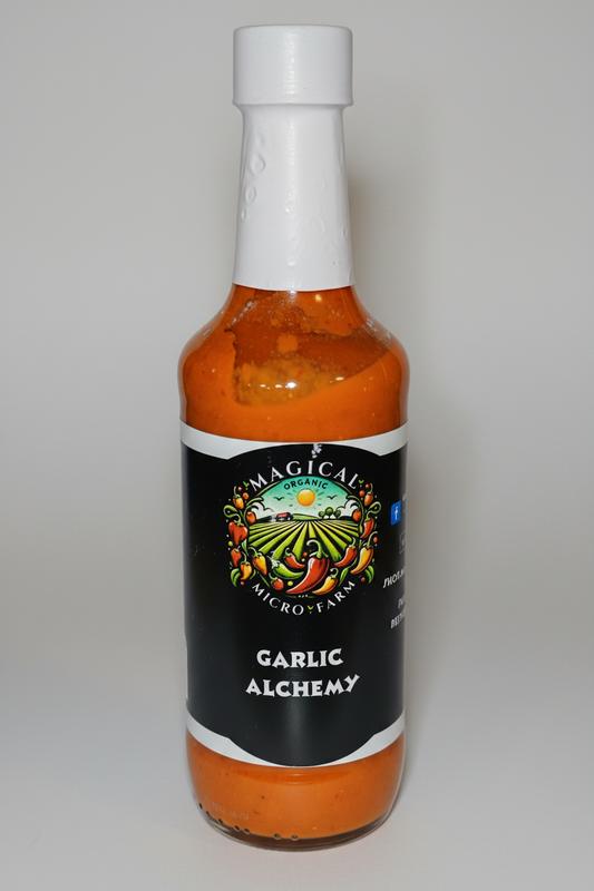 A bottle of Garlic Alchemy hot sauce by Omagical Alchemy, featuring a black label with a colorful farm illustration and white cap, filled with orange-red sauce, isolated on a white background.