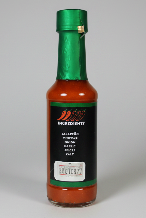 A bottle of hot sauce with a green foil neck wrap, a black label reading 'Ingredients' and 'Jalapeño Vinegar Onion Garlic Spices Salt', and orange-red sauce visible through the clear bottle, isolated on a white background.